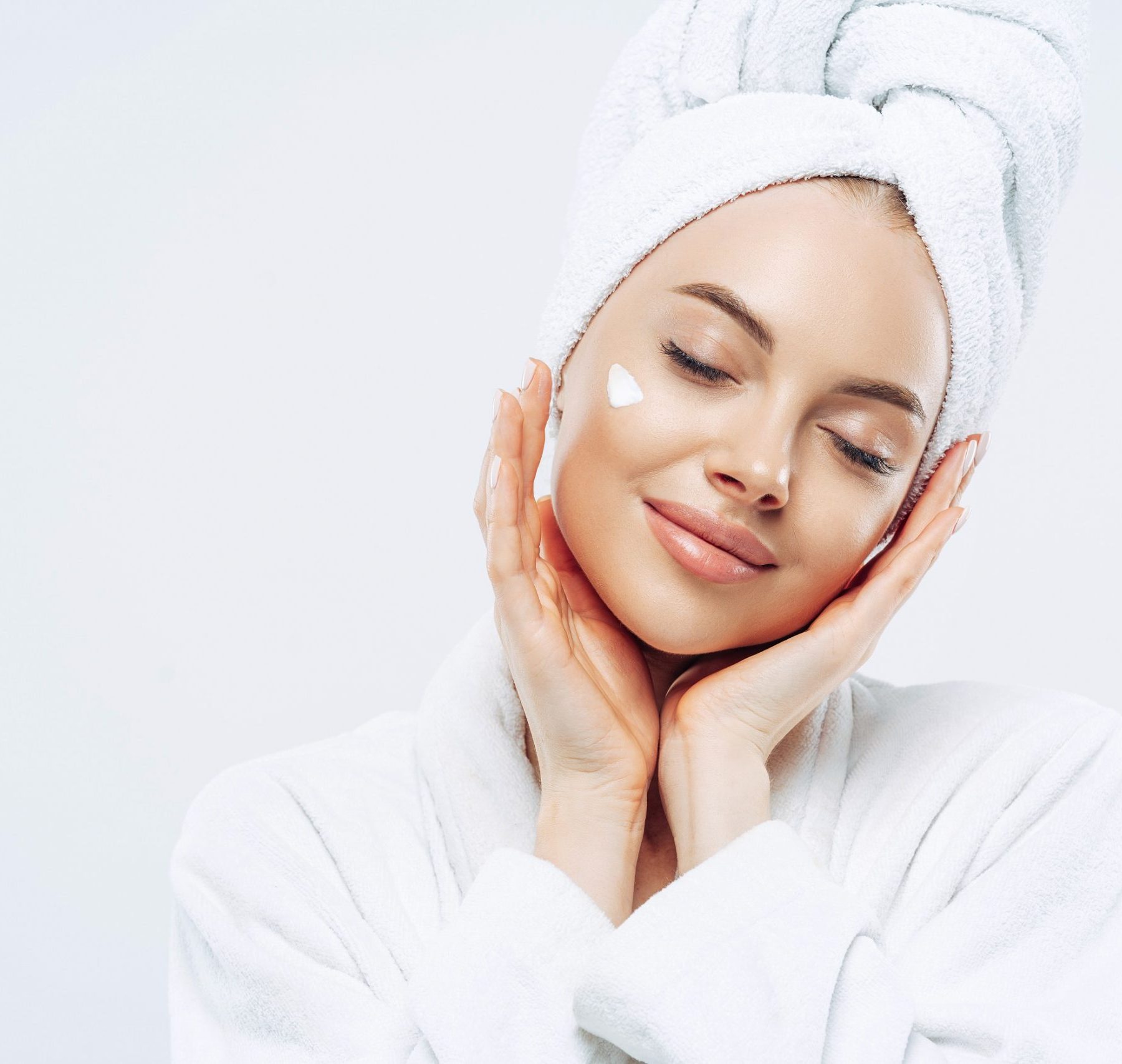 Studio shot of relaxed beautiful woman with closed eyes tilts head, touches skin gently, applies face cream, wears bath towel on head after taking spa procedures, enjoys facial treatments, body care Studio shot of relaxed beautiful woman with closed eyes tilts head, touches skin gently, applies face cream, wears bath towel on head after taking spa procedures, enjoys facial treatments, body care